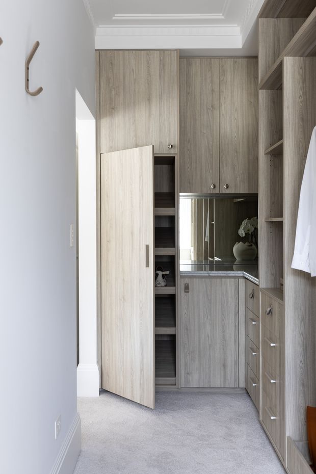 A compact, well-organised walk-in wardrobe with floor-to-ceiling light wood-grain cabinetry, open shelving, drawers, hanging storage and a central mirrored vanity or dressing counter. The room has soft grey carpet, pale grey walls, white decorative ceiling moulding and subtle brushed metal hardware. A partially open tall cabinet reveals additional shelving, while clothing and accessories indicate dressing-room use. The space is bright, refined and highly tailored.