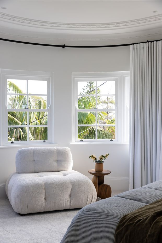 A bright, elegant bedroom corner with two white-paned windows overlooking palm foliage, ornate decorative ceiling cornices, full-height white curtains on a black curtain rod, plush off-white boucle lounge chair, small timber side table, and the edge of a neatly made bed with a grey ribbed cover and dark throw. The room has a soft, refined, neutral palette and abundant natural light.