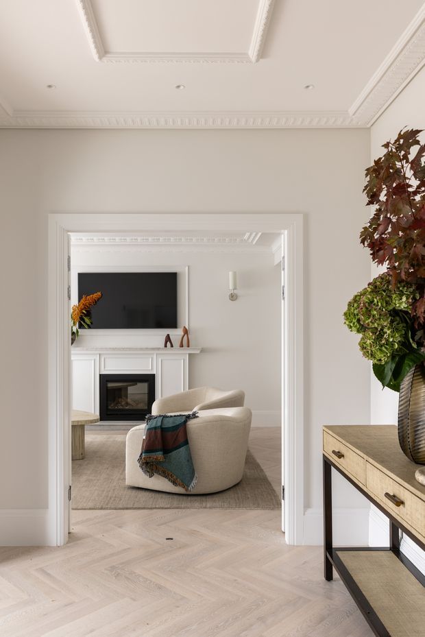 A refined residential interior view through a wide trimmed doorway into a living room. The foreground has pale herringbone timber flooring, warm off-white walls, elaborate decorative ceiling cornices and a console table with a large floral arrangement. Beyond the doorway is a softly furnished sitting room with a white traditional fireplace, black television, upholstered armchair, neutral rug and a wall sconce. The overall scheme is light, calm and elegant with strong classical detailing.