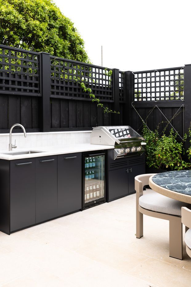 A compact, refined outdoor entertaining area with a built-in black outdoor kitchen, white tiled splashback and benchtop, sink, under-counter drinks fridge, and stainless-steel barbecue. The space is enclosed by dark timber fencing with decorative lattice screens and greenery. A round dining table with upholstered chairs occupies the foreground, and pale stone-look paving creates a bright contrast with the dark cabinetry.