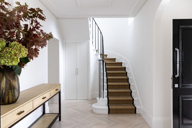 Bright, refined residential entry hall featuring a curved staircase with dark timber treads, a textured runner, and slim black metal balustrade. White paneled walls and ceiling, built-in storage, pale herringbone flooring, a timber console table, and a bold black entry door create a high-contrast, elegant interior.