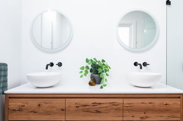 Bright contemporary bathroom vanity featuring a wide natural timber floating cabinet with three drawers, a white benchtop, two white vessel basins, matte black wall-mounted taps, and twin round mirrors. A small potted plant adds greenery between the basins.