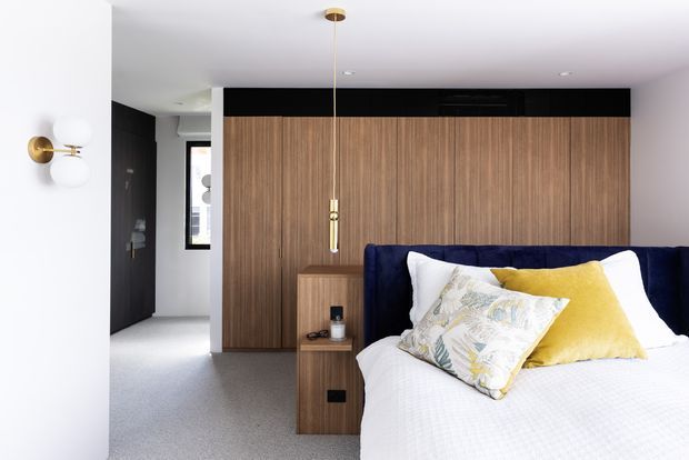 Contemporary bedroom with a partially visible ensuite or hallway area, featuring a full-width timber wardrobe and headboard wall, dark charcoal side walls, integrated bedside joinery, navy upholstered bed, white bedding, and mustard and patterned accent cushions. Brass wall and pendant lighting add warm highlights to the otherwise restrained palette.