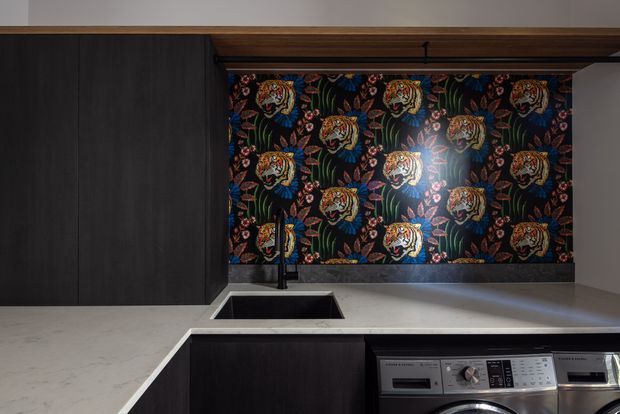 A close-up view of a contemporary kitchen and integrated laundry area. Dark timber cabinetry contrasts with a pale stone benchtop, while a black sink and tap sit beneath a dramatic patterned backsplash featuring tigers and foliage. Front-loading laundry appliances are integrated below the bench on the right. The space has a moody, refined character with warm timber detailing and bold decorative patterning.