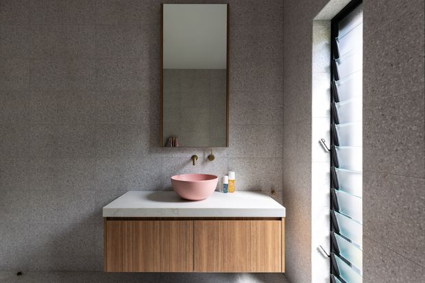 A compact contemporary bathroom vanity area with a wall-mounted timber-grain cabinet, white benchtop, pink vessel basin, brass tapware, and a tall rectangular mirror. The walls and floor are clad in dark grey textured tile or stone, with a louvred window bringing in natural light.