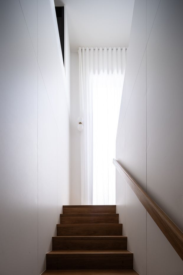 A narrow, minimalist stairway with dark timber treads and matching timber handrail set against crisp white walls. The stairs lead upward toward a tall, softly curtained window, creating a bright vertical focal point and strong contrast between the warm wood and white architectural surfaces.