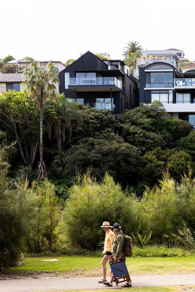 Exterior streetscape view of modern multi-storey residential homes set among dense subtropical landscaping. The central dark-clad dwelling has a steep gable roof, projecting balcony, expansive glazing and a strong black-and-glass facade, while neighbouring contemporary homes are visible on either side. A landscaped pedestrian path and lawn occupy the foreground.