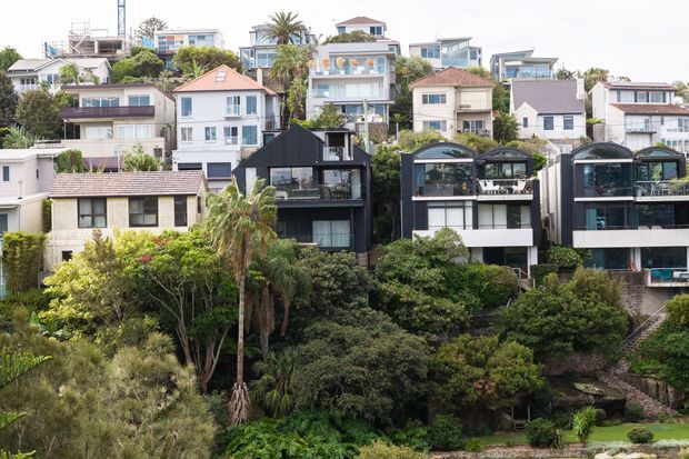 Wide exterior view of a dense hillside suburban neighbourhood with a mix of contemporary and traditional multi-storey homes, lush established vegetation, balconies, terraces, and varied roof forms. Several dark-clad modern residences stand prominently in the foreground, while lighter homes are arranged up the slope behind them.