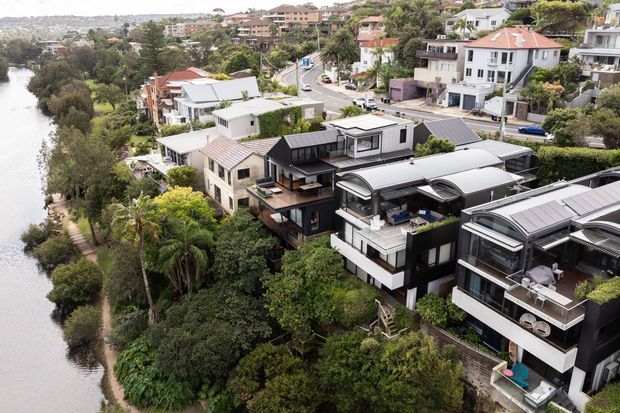 Aerial view of a dense residential neighbourhood beside a narrow waterfront or river. The foreground is dominated by contemporary multi-level homes with dark cladding, white rendered walls, extensive glazing, rooftop terraces, and curved metal roof forms. Lush established vegetation separates the properties from the water, while additional detached homes and apartment-style residences climb the opposite slope in the background.