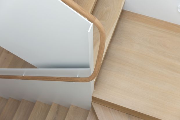 Close-up architectural view of a modern timber staircase and landing. Pale oak-toned stair treads and landing flooring contrast with smooth white walls, while a rounded timber handrail follows the stair edge around the landing. The image emphasizes clean joinery, warm natural wood, and a minimalist stair design.