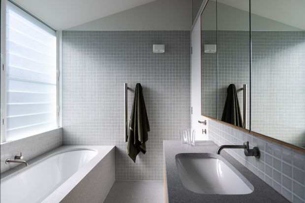 A contemporary bathroom with a freestanding white bathtub beside a large louvred window, a wall-mounted grey mosaic tile finish, and a grey stone vanity benchtop with an integrated basin. A large mirror reflects the towel rail and tiled walls. The room has clean lines, restrained grey-and-white colours, and abundant natural light.
