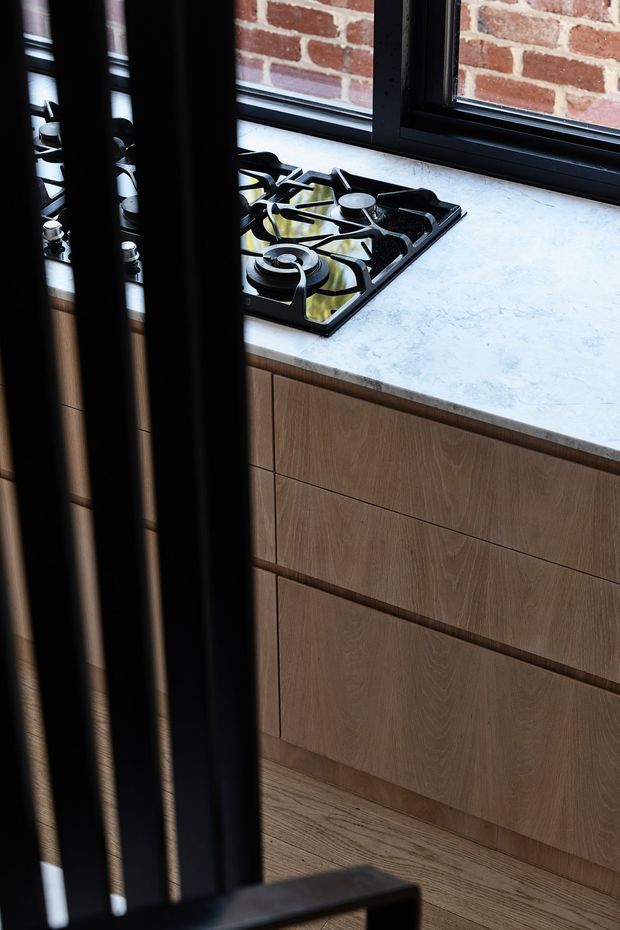 Close-up view of a contemporary kitchen, focused on a black gas cooktop set into a pale stone countertop above warm timber-grain cabinetry. Black-framed windows reveal an exposed red-brick wall outside, while dark vertical slats partially screen the foreground.