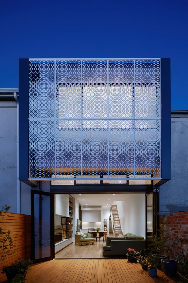 Twilight view of a contemporary multi-level urban residence with a striking illuminated patterned metal screen enclosing the upper facade. Large glazed bi-fold or sliding doors open from the bright ground-floor living area onto a timber deck, with built-in shelving, sofas, a dining table, and a stair visible inside. Existing rendered and exposed brick walls frame the extension, while potted plants and a timber boundary screen surround the outdoor deck.
