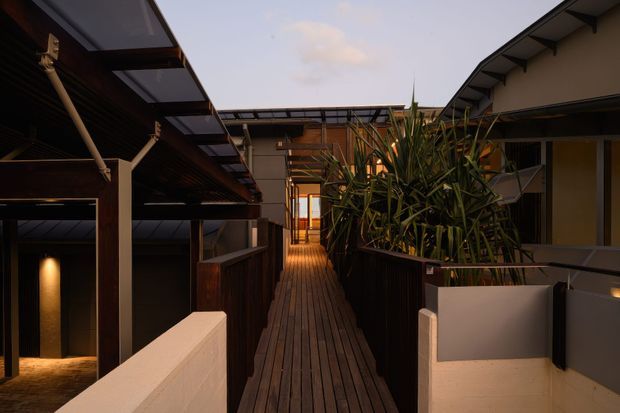 A narrow elevated timber walkway passes through a modern multi-level building complex, with dark timber decking, timber posts and beams, translucent roof awnings, smooth rendered walls, metal balustrades, and dense architectural planting in raised planters. Warm interior lighting contrasts with the dusky sky, creating a sheltered courtyard-like circulation space.