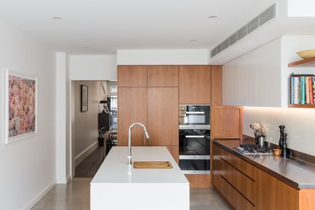 Bright contemporary kitchen with a large white island, integrated sink and timber cabinetry. The room includes a dark stone benchtop, built-in stainless-steel appliances, under-cabinet lighting, concealed ventilation, and a compact dining or hallway area beyond the kitchen. White walls and ceilings create a clean, refined interior.