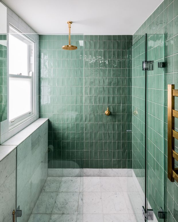 A bright, refined bathroom shower enclosure lined with glossy green rectangular tiles. The space features a large white-framed window, pale marble or stone flooring and lower wall surfaces, a frameless glass shower screen, and a brass/gold rainfall shower fixture with matching controls. A timber-toned heated towel rail is visible at the right edge.