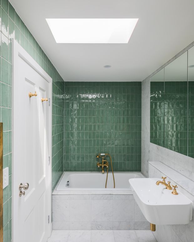 A compact, elegant bathroom with glossy deep green square wall tiles, white marble-look wall and floor surfaces, a built-in bathtub with brass fixtures, and a wall-mounted white basin with vintage-style brass taps. A large mirrored cabinet and rectangular skylight add brightness and reflect the tiled interior.