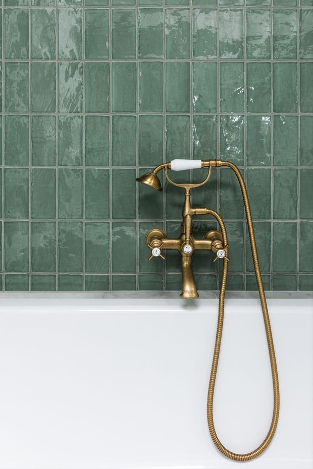 Close-up view of a bathtub shower fixture against a full wall of glossy, irregular green ceramic tiles. The fixture has an antique brass finish, exposed mixer controls, a handheld shower hose, and a white freestanding or built-in tub below. The composition is focused on bathroom fittings, tilework, and contrasting brass and green finishes.