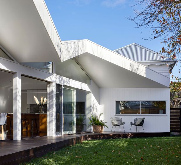 A bright contemporary white-clad residential extension viewed from the garden. The composition features angular roof forms, a deep covered deck, large glazed openings, and a partially visible warm timber kitchen inside. A small outdoor seating area sits beside the lawn, with mature tree shadows across the facade.