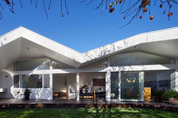 A single-storey modern residence viewed from the garden, with a white angular flat-roof facade, deep covered verandah, extensive glazing, and a central recessed entry. The home opens onto a timber deck, with outdoor planting and strong tree-shadow patterns across the walls. A clear blue sky and sparse branches frame the upper edge.