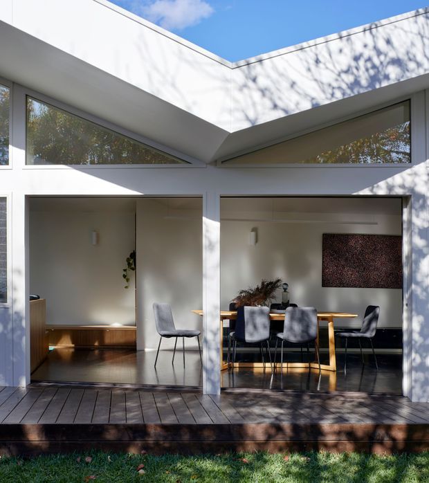 A bright modern courtyard-facing residential facade with white rendered walls, a flat roof, high clerestory glazing, and two large open living spaces overlooking a timber deck. The left room appears to be a living or sitting area, while the right room contains a dining table and chairs. Strong sunlight and tree shadows create a crisp indoor-outdoor atmosphere.