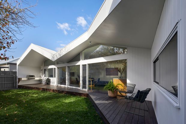 Exterior view of a contemporary single-storey residence with a dramatic angular roof and deep overhangs. White vertical cladding, extensive floor-to-ceiling glazing, and a dark timber deck frame a compact landscaped courtyard. The interior living area is visible through the windows, while an outdoor dining setting occupies the deck.