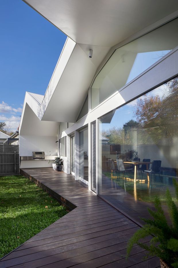Exterior view of a contemporary white-clad home with strong geometric roof overhangs, extensive floor-to-ceiling glazing, and a dark timber deck wrapping around the side. The deck includes an outdoor barbecue area, while a dining space is visible through the glass. The image is photographed in a sunny suburban garden setting with a clean, modern architectural composition.