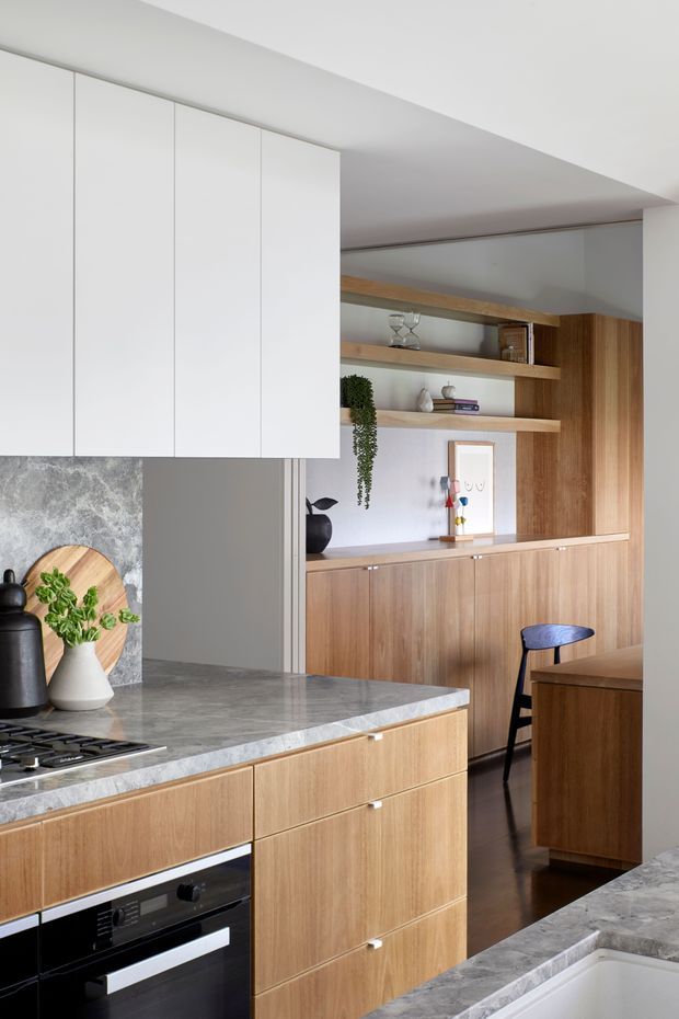 A refined contemporary kitchen with extensive warm timber cabinetry, white overhead cupboards, and a grey veined stone benchtop. Integrated appliances and a gas cooktop occupy the foreground island, while a timber-lined wall with open display shelving and cabinetry forms the background. Minimal decor, sculptural ceramics, greenery, and a dark blue bar stool add restrained colour and character.
