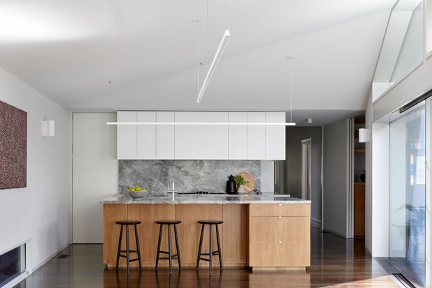 A bright, contemporary kitchen with a central timber island featuring three black bar stools, white overhead cabinetry, and a dark veined stone splashback and benchtop. The room has polished dark timber flooring, white walls and ceiling, minimalist linear pendant lighting, and extensive glazing along the right side.