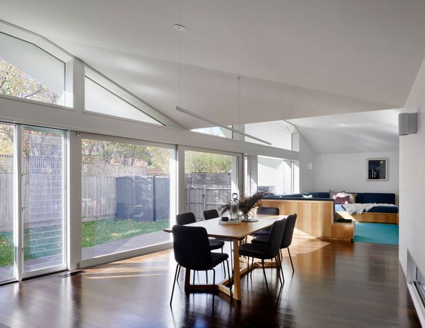 Bright contemporary open-plan living and dining area with extensive floor-to-ceiling glazing, a pitched/sloping ceiling with clerestory windows, polished timber flooring, and a compact dining table with dark upholstered chairs. A raised timber built-in platform and blue upholstered seating occupy the rear, while the glazing opens views to a fenced garden.
