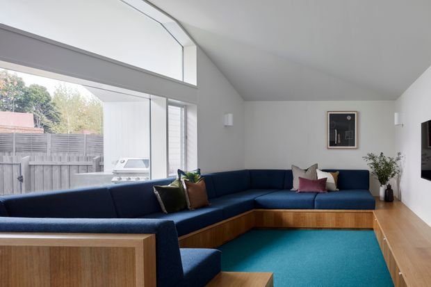 A bright contemporary living room with a large navy-blue L-shaped built-in sectional sofa, warm timber panelled seating base and cabinetry, turquoise carpet, white walls, and extensive corner glazing. The glazing opens visually to an outdoor barbecue area and fenced garden, while minimalist artwork and a potted plant add restrained decoration.