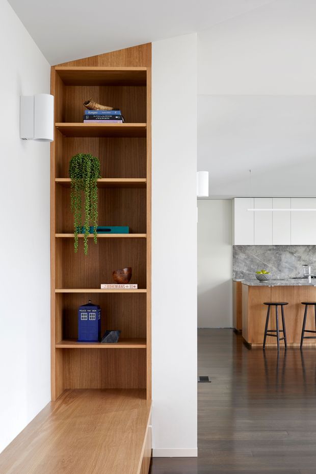 Bright contemporary open-plan interior featuring a tall built-in timber bookshelf integrated into a white wall, with a timber ledge or desk surface below. Through the opening, a modern kitchen is visible with white overhead cabinetry, warm timber island cabinetry, a grey stone-look splashback, and dark timber flooring. Minimal cylindrical wall and pendant lighting complete the clean, restrained design.