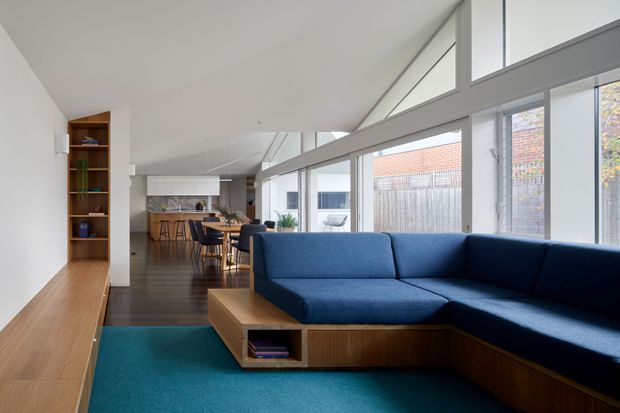 Bright open-plan living area with a large built-in deep blue sectional sofa, warm timber media/storage joinery and coffee table, and a vivid teal rug. The space extends toward a contemporary kitchen and dining area, with extensive floor-to-ceiling glazing and high clerestory windows providing abundant natural light. Clean white surfaces and dark timber flooring create a modern, airy interior.