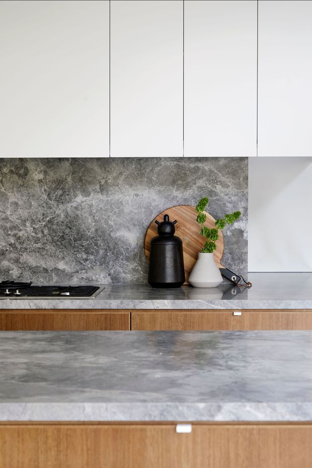 Close-up view of a refined contemporary kitchen featuring white flat-panel overhead cabinetry, a dramatic grey veined stone splashback and benchtops, a black gas cooktop, and warm timber-grain lower cabinetry. Minimal styling includes a round timber board, black vessel, white vase, and greenery.