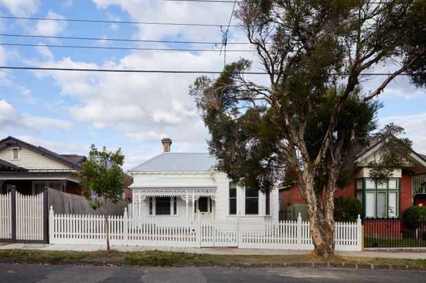 Street-facing view of a single-storey heritage cottage with an ornate white-painted verandah, decorative fretwork, corrugated metal roof, tall sash-style windows, and a white picket fence. Mature street trees and neighbouring period homes frame the suburban residential setting.