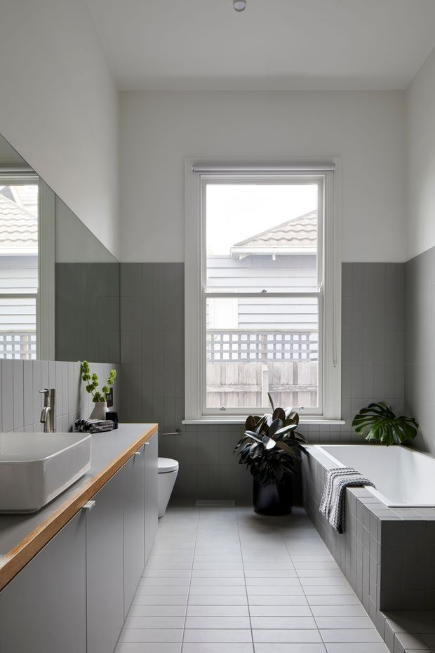 A bright, narrow contemporary bathroom with a freestanding-style white basin on timber-edged grey vanity cabinetry, a toilet, and a tiled built-in bathtub. Grey square wall tiles extend to mid-height, with pale floor tiles and white upper walls. A large white-trimmed window brings in natural light, while potted plants and folded towels add warmth and texture.