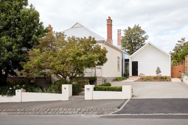 Street-facing view of a traditional light-weatherboard house with multiple red brick chimneys, a steep gabled roof, landscaped front garden, low white boundary wall, and a modern detached white garage or studio accessed by a broad concrete driveway.