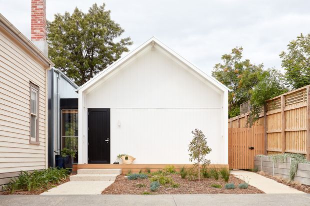Exterior view of a compact white gable-roofed detached structure in a landscaped courtyard or side yard. The facade has vertical white cladding, a black entry door, simple white trim, and a small timber-toned base. It is positioned beside an existing light weatherboard building, with timber privacy fencing, concrete block retaining walls, concrete paths, and low-maintenance planting.