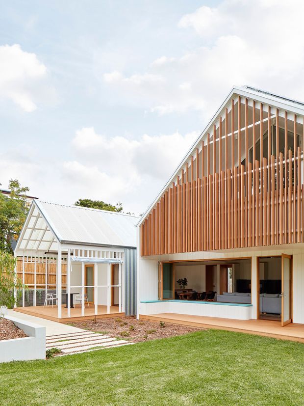 Exterior view of a contemporary residential building with a steep gabled roof, white metal cladding, extensive vertical timber screening, and large glazed openings. A smaller matching pavilion with a covered outdoor deck sits beside the main structure, connected by a landscaped lawn and stepping-stone path. The image highlights the building facade, roof forms, outdoor living areas, and timber sun-screening.