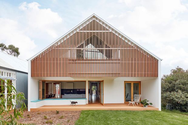 Front exterior view of a contemporary two-storey gable-roof house. The facade combines white vertical cladding with a prominent upper-level screen of closely spaced warm timber slats, creating a semi-transparent gabled balcony or upper living area. Large glazed openings and timber-framed doors connect the ground floor to a narrow timber deck and landscaped lawn. The design features a strong symmetrical gable composition and a screened upper facade.