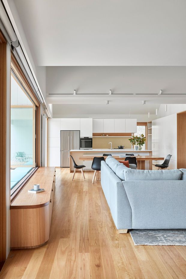 Bright open-plan contemporary interior combining a living room, dining area and kitchen. The space features extensive timber-framed glazing along the left side, wide light timber flooring, a pale blue-grey sectional sofa, a timber dining table, white flat-panel kitchen cabinetry, a timber-toned island and integrated appliances. Minimal linear ceiling lighting and clean built-in joinery create an airy, refined appearance.