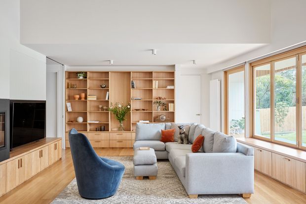 Bright contemporary living room with a large light-grey sectional sofa, deep blue accent armchair, timber built-in media unit and full-height shelving, and a broad timber-framed window seat beside expansive glazing. The room has pale timber flooring, white walls and ceiling, recessed lighting, and a neutral palette with rust-coloured cushions and natural decorative accents.