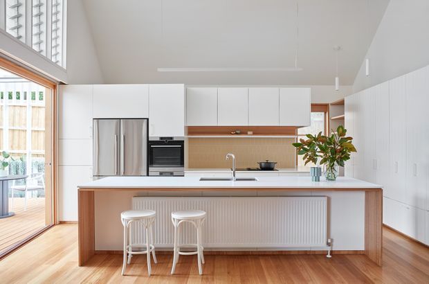 Bright contemporary kitchen with a large white island and integrated seating, flat-panel white cabinetry, stainless-steel refrigerator and oven, warm timber detailing, a textured tan backsplash, and timber flooring. Full-height glazing and a glazed door bring in abundant natural light, while a high angled ceiling creates an airy feel.