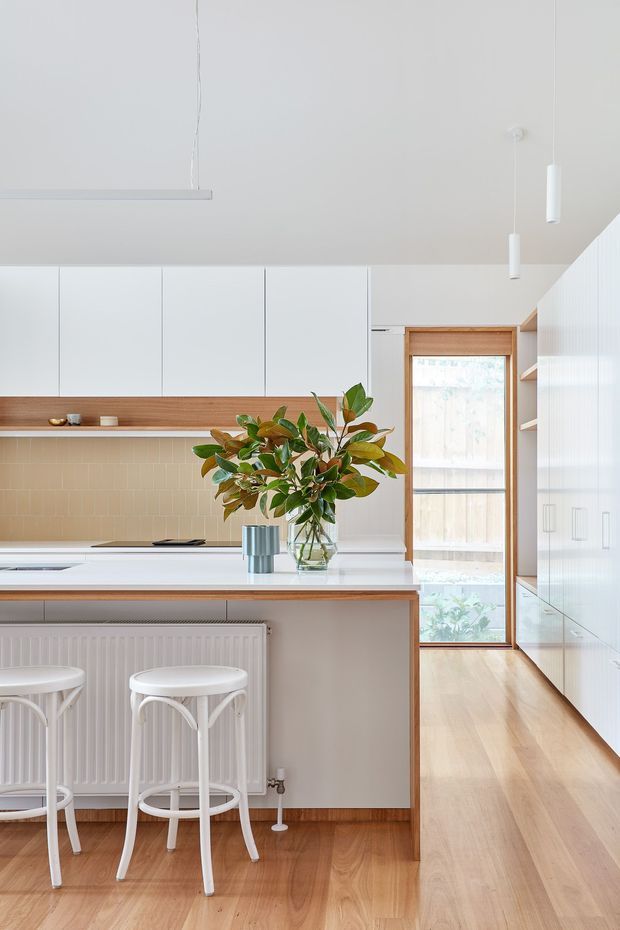 Bright, compact contemporary kitchen with white flat-panel cabinetry, a pale stone island benchtop, warm timber flooring and timber accents, and a beige tiled splashback. Two white bar stools sit at the island, while a vase of leafy branches forms a natural centrepiece. A tall timber-framed glazed door or window brings in daylight, and slim white pendant lights hang from the high ceiling.