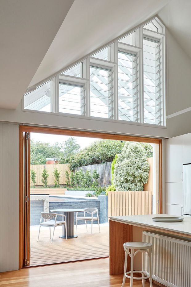 Bright contemporary kitchen opening onto a timber deck and landscaped outdoor entertaining area with a pool. The kitchen has white cabinetry, a pale benchtop, timber joinery, a peninsula with a bar stool, and a large timber-framed glazed opening. Above it is a dramatic bank of high clerestory windows with louvres and triangular glazing that brings in abundant natural light.
