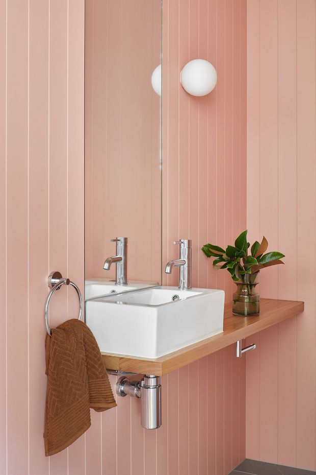 A compact contemporary bathroom vanity featuring a white vessel basin, chrome wall-mounted tapware and exposed plumbing, set against a blush-pink vertically panelled wall. A large mirrored cabinet or mirror spans the vanity, with a round white wall light, timber benchtop, chrome towel ring, brown hand towel, and a small potted plant adding warmth.