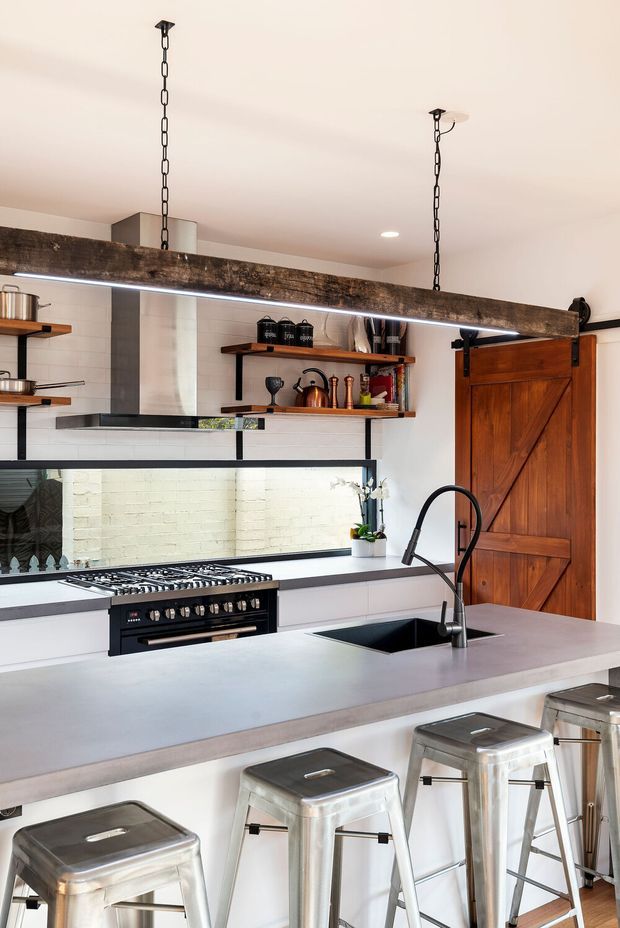 Bright industrial-style kitchen with a large pale stone island, integrated dark sink and gooseneck tap, professional freestanding range, open timber shelving, white subway-tile splashback, and a rustic timber barn door. Exposed dark timber beam and hanging linear lighting add character, while metal bar stools provide seating at the island.