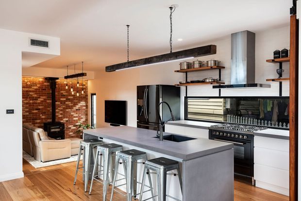 Open-plan contemporary kitchen with a large grey island and four metal bar stools, black appliances, white cabinetry, open timber shelving, and a prominent stainless-steel range cooker. A brick feature wall, wood-burning stove, and living area are visible beyond the kitchen. Two suspended timber-and-metal pendant lights add an industrial accent.