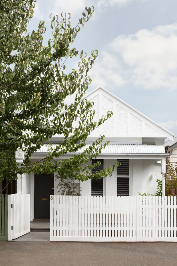 Street-facing view of a compact white-painted house with a prominent gabled front facade, textured wall panels, a corrugated metal awning, louvred windows, and a white vertical-slat front fence and gate. Mature leafy planting partially screens the facade, with a paved street in the foreground and a bright sky above.