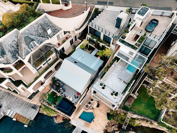 Aerial oblique view of a dense waterfront residential neighbourhood with several architecturally distinctive homes, rooftop terraces, landscaped courtyards, timber decks, swimming pools, and direct water frontage. The buildings include varied roof forms, pale rendered facades, metal roofing, extensive glazing, and multiple outdoor entertaining areas.